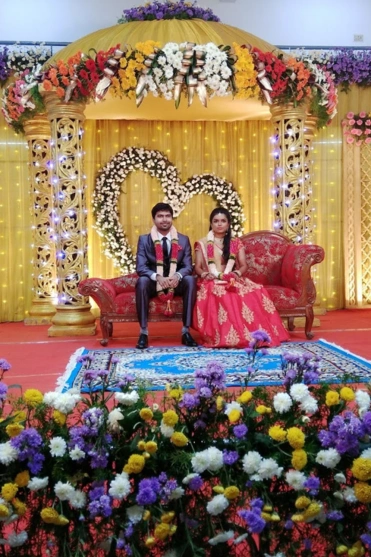 Two individuals are seated on a sofa at a wedding stage decorated with flowers & a golden backdrop.
