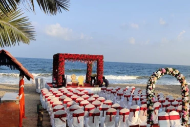 Beach wedding setup with floral arch, white chairs and red accents, by the sea under a clear sky.